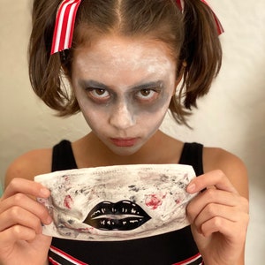 May include: A white fabric face mask with a black and red lips design. The mask is stained with red and black paint, giving it a spooky look.