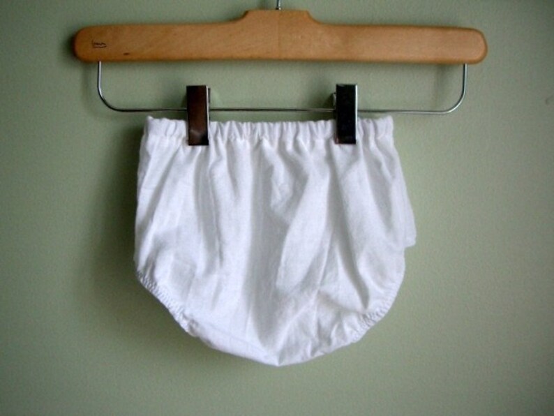 May include: White cotton baby bloomers hanging on a wooden hanger with silver clips.
