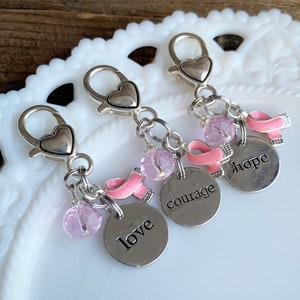 May include: Three silver keychains with heart-shaped clasps and charms. Each keychain has a pink ribbon charm, a pink bead, and a silver disc with the words "love", "courage", and "hope". The keychains are displayed on a white, decorative plate.