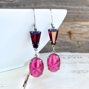 May include: A pair of dangle earrings with a vintage aesthetic. Each earring features a deep red, geometric top and a pink, oval-shaped bottom. The earrings are connected by a silver-toned metal chain and hook.