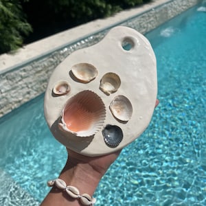 Op de afbeelding: Witte keramische serveerschaal met een gat in de handgreep, met een verzameling schelpen in verschillende maten en kleuren. De schaal wordt vastgehouden door een hand met een witte schelpenarmband.