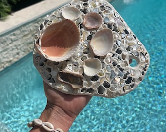 May include: A mosaic stone art piece featuring a variety of seashells and small pebbles in shades of white, beige, and gray. The piece is held up against a backdrop of a swimming pool.