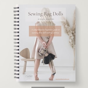 Pode incluir: Um livro com espiral intitulado "Sewing Rag Dolls" de Dainy Sawatzky. A capa apresenta uma criança segurando uma boneca de pano, com o texto "A comprehensive beginners guide to starting your doll making journey."