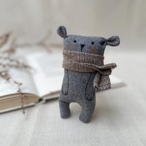 May include: A handmade gray stuffed animal bear with a plaid scarf. The bear has small ears, rosy cheeks, and black stitched details. The scarf is a mix of brown, red, and gray. The bear is standing in front of an open book.