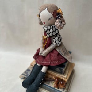 May include: A handmade fabric doll with brown hair, wearing a dark red dress, a beige coat, and a black and white polka dot collar. The doll features embroidered facial details, including closed eyes and red lips, and is adorned with small fabric flowers.