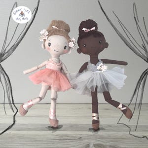 Könnte beinhalten: Zwei handgefertigte Ballerina-Puppen. Eine Puppe hat helle Haut, einen rosa Tüllrock und einen Blumenkranz. Die andere Puppe hat dunkle Haut, einen weißen Tüllrock und einen Dutt. Beide Puppen befinden sich in Balletthaltungen, mit Ballettschuhen und Bändern.