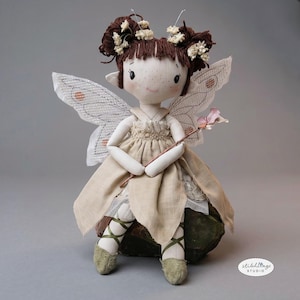 Op de afbeelding: Een handgemaakte feeënpop met bruin haar versierd met kleine witte bloemen. De pop draagt een beige jurk, kanten vleugels en groene balletschoenen. Ze houdt een roze toverstaf vast en zit op een groene steen.