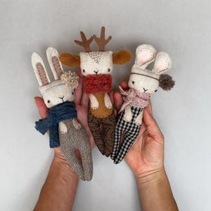 Könnte beinhalten: Drei handgefertigte Stoffpuppen: ein Rentier mit Geweih und rotem Schal und zwei Hasen mit Hüten und Schals. Die Hasen haben lange Ohren und tragen blaue und rosa Schals. Die Puppen haben gemusterte Hosen.