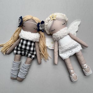 May include: Two handmade fabric dolls with embroidered facial features. The left doll has blonde yarn hair, a black and white gingham dress, a white textured collar, and grey striped leg warmers. The right doll has white hair, a white lace dress, white wings, and a yellow star hair accessory.