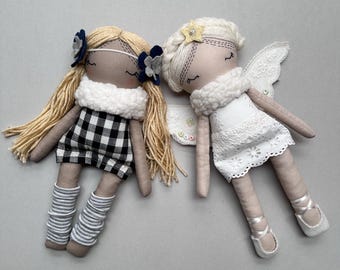 May include: Two handmade fabric dolls with embroidered facial features. The left doll has blonde yarn hair, a black and white gingham dress, a white textured collar, and grey striped leg warmers. The right doll has white hair, a white lace dress, white wings, and a yellow star hair accessory.