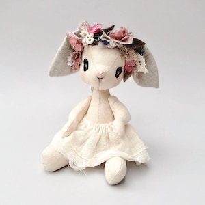 Puede incluir: Una muñeca de peluche de conejo hecha a mano con un cuerpo color crema, orejas grises y una falda blanca. El conejo lleva una corona floral con detalles en rosa y marrón. La muñeca tiene ojos negros y está sentada.