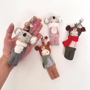 以下が含まれることがあります： 動物のフードを被ったキャラクターのぬいぐるみキーホルダー4点。グレーのコアラ、ライトグレーの生き物、緑のマフラーをした茶色のクマ、赤のマフラーをした茶色のクマが含まれています。それぞれに金属製のリングと小さな星のチャームが付いています。