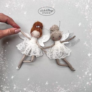 May include: Two handmade fairy dolls in white lace dresses and wings. One doll has red hair, the other has tan hair. The dolls are holding hands. The image has the text "stitchlings STUDIO".
