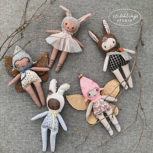 May include: Five handmade fabric dolls, including fairies and bunnies, are arranged on a grey surface. The dolls feature detailed stitching, with some wearing tutus, wings, and hats. The dolls are in various colours, including pink, brown, and blue.