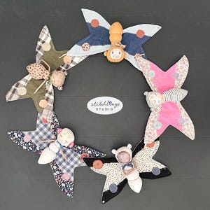 Op de afbeelding: Een cirkelvormige opstelling van vijf vlindervormige stoffen poppen, elk met een uniek textielpatroon en kleurenschema. De poppen hebben geborduurde gezichten en zijn geplaatst rond een centraal wit ovaal label met de tekst stitchlings STUDIO.