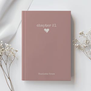 May include: A dusty rose-colored book with the text "chapter 21" and a heart symbol. The name "Charlotte Grace" is printed at the bottom. The book is surrounded by white flowers and a white cloth.