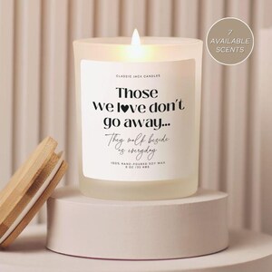 May include: A lit, frosted glass candle with a wooden lid, sitting on a white pedestal. The candle has text that reads "Those we love don't go away... They walk beside us everyday." The candle is 8 oz and made of 100% hand-poured soy wax.