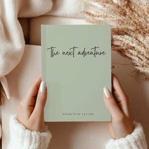 May include: A sage green journal with the words "The next adventure" in elegant script. The author's name, Charlotte Taylor, is printed at the bottom. The journal is held by a person wearing a white sweater.