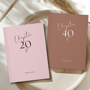 May include: Two pink and brown hardcover journals with the text "Chapter 20" and "Chapter 40" on the front covers. The journals have a simple floral design and the name "Amelia Hart" printed on the bottom of each cover.
