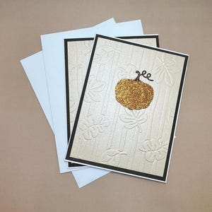 May include: Greeting cards with a fall theme. The cards feature a gold glitter pumpkin and embossed leaf designs on a cream background. The cards have a black border and are paired with light blue envelopes.