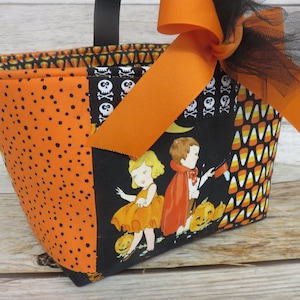 May include: A Halloween fabric tote bag in orange and black. The bag has a black handle and a large orange bow. The fabric is patterned with polka dots, candy corn, and vintage Halloween illustrations, creating a festive look.