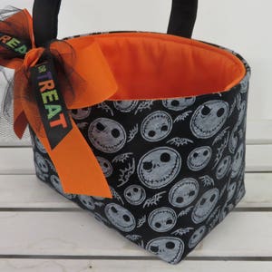 Puede incluir: Un bolso tote con tem&aacute;tica de Halloween con un exterior negro estampado con caras de Jack Skellington. El bolso tiene un interior naranja y est&aacute; adornado con un lazo naranja y una cinta negra que dice "TREAT". Las asas negras completan el dise&ntilde;o.