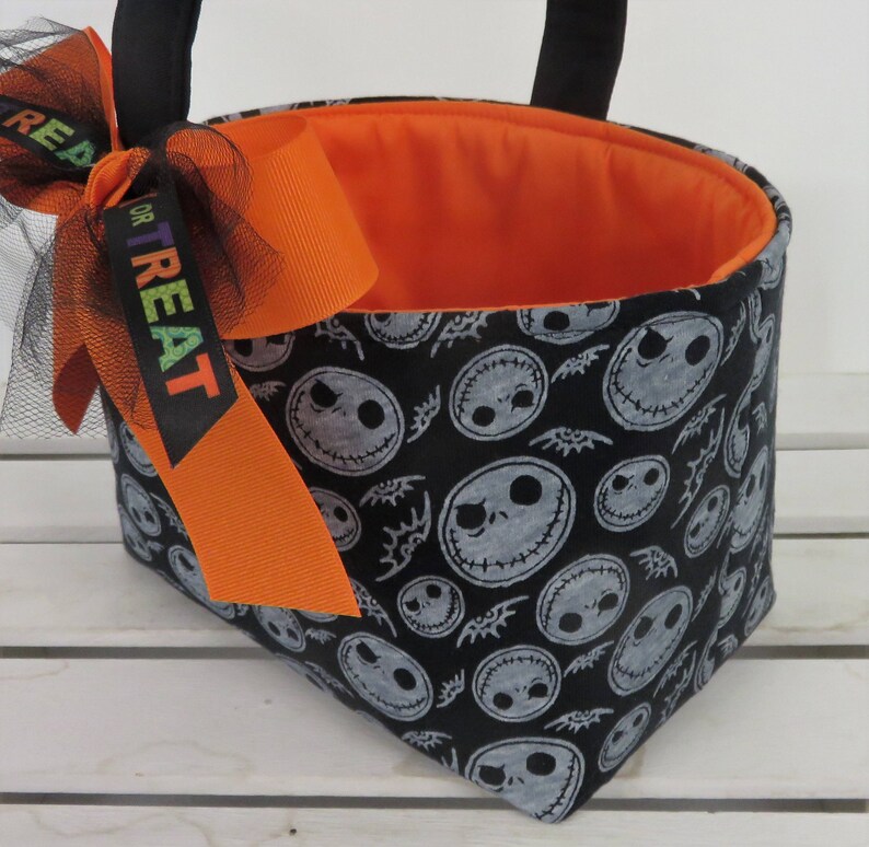Puede incluir: Un bolso tote con tem&aacute;tica de Halloween con un exterior negro estampado con caras de Jack Skellington. El bolso tiene un interior naranja y est&aacute; adornado con un lazo naranja y una cinta negra que dice "TREAT". Las asas negras completan el dise&ntilde;o.