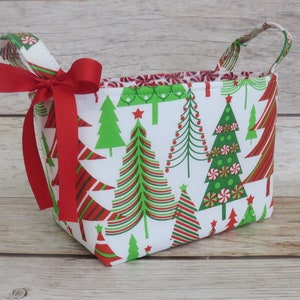 Puede incluir: Una cesta rectangular de tela con un estampado de árboles de Navidad en verde y rojo sobre fondo blanco. La cesta tiene dos asas y una cinta roja atada con un lazo. El ribete interior presenta un patrón de bastones de caramelo.
