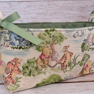 May include: A green fabric storage basket with a Winnie the Pooh and friends print. The basket has two green fabric handles and a green satin bow.