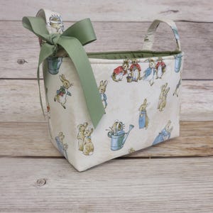 May include: A fabric basket with a Peter Rabbit print, featuring bunnies in various poses with watering cans and other garden elements. The basket has a green interior and a sage green bow. The handles are made of the same fabric as the basket.