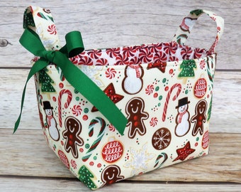 May include: A white fabric storage basket with green handles and a green bow. The basket is decorated with a Christmas theme featuring gingerbread men, candy canes, snowmen, and snowflakes.