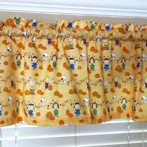 May include: A yellow valance with a pattern of Peanuts characters and pumpkins. The valance is made of fabric and has a gathered look.
