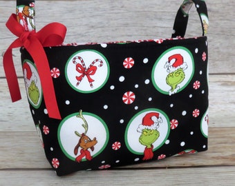 May include: A rectangular fabric storage basket with a black background and a repeating pattern of Grinch characters, candy canes, and snowflakes. The basket has red handles and a red bow.