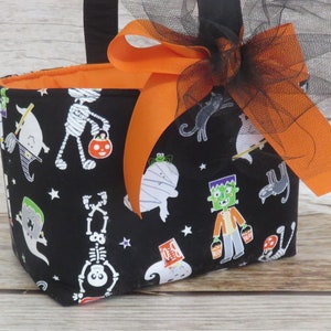 May include: A Halloween-themed fabric basket with a black exterior featuring white ghosts, skeletons, and other spooky characters. The basket has an orange interior and is adorned with an orange ribbon and black tulle bow.