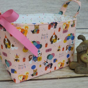 May include: A pink fabric storage basket with a dog-themed print, a pink bow, and handles. The basket is decorated with cartoon dogs, balloons, and gift boxes. A small dog figurine sits beside the basket.