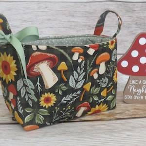 May include: A green fabric storage basket with a mushroom and sunflower pattern. The basket has two fabric handles and a green ribbon tied around the handles. The basket is sitting on a wooden surface next to a red and white mushroom-shaped wooden sign that says "Like a good neighbour stay over there."