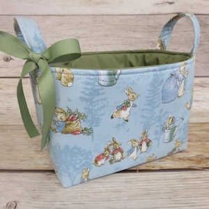 May include: A rectangular fabric basket with handles and a green bow. The light blue fabric features a repeating pattern of rabbits in a forest setting. The interior lining is green.