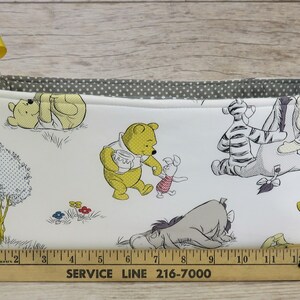 May include: A white fabric storage basket with yellow handles and a Winnie the Pooh print. The basket features Winnie the Pooh, Piglet, Tigger, Eeyore, and other characters from the Disney cartoon. The basket is 15 inches long.