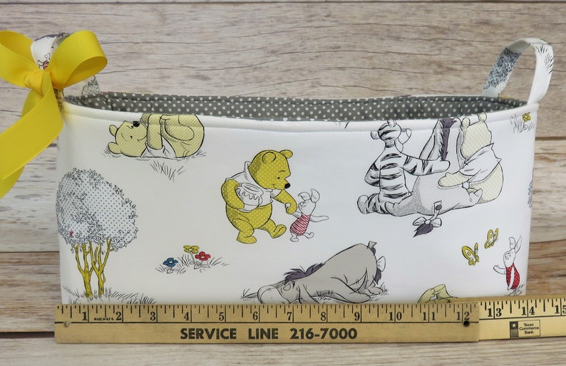 May include: A white fabric storage basket with yellow handles and a Winnie the Pooh print. The basket features Winnie the Pooh, Piglet, Tigger, Eeyore, and other characters from the Disney cartoon. The basket is 15 inches long.