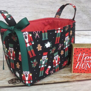 May include: A black fabric storage basket with a red and white polka dot lining. The basket is decorated with a pattern of nutcracker figures and gingerbread men. The basket has two green fabric handles and a green ribbon bow. The basket is sitting on a wooden surface next to a wooden block with the text "Home Sweet Home" in white.