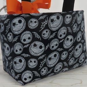 Puede incluir: Bolso tote de tela negra con un patr&oacute;n repetido de caras de Jack Skellington. El bolso tiene asas negras y est&aacute; decorado con un lazo naranja y una cinta plateada.