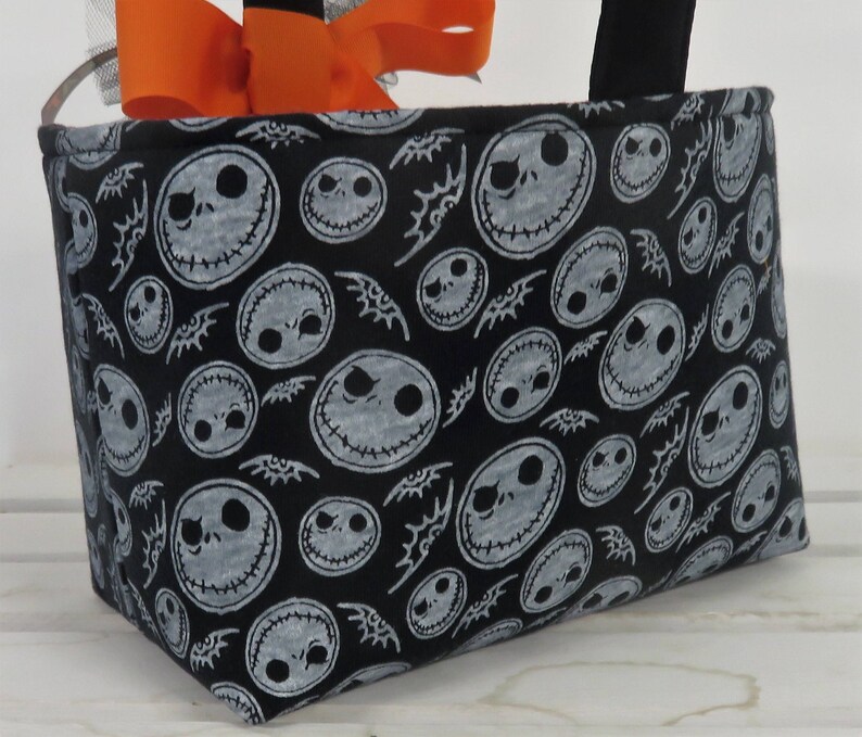 Puede incluir: Bolso tote de tela negra con un patr&oacute;n repetido de caras de Jack Skellington. El bolso tiene asas negras y est&aacute; decorado con un lazo naranja y una cinta plateada.