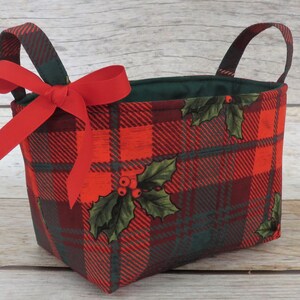 May include: A rectangular fabric basket with a red plaid pattern and green accents. The basket features a red bow and holly leaf and berry details. It has two handles and a green interior.