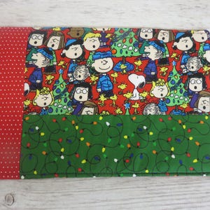 May include: A rectangular fabric mat featuring a red side panel with white polka dots. The main section displays a Peanuts holiday print with characters singing around Christmas trees, while the bottom section is green with string lights.