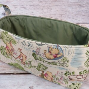 May include: A Winnie the Pooh themed fabric storage basket with green lining and two handles. The basket features a white background with illustrations of Winnie the Pooh, Piglet, Tigger, and Eeyore.