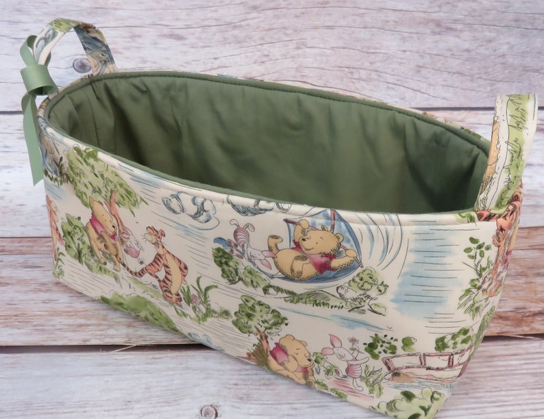 May include: A Winnie the Pooh themed fabric storage basket with green lining and two handles. The basket features a white background with illustrations of Winnie the Pooh, Piglet, Tigger, and Eeyore.