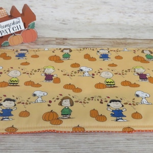 May include: A yellow fabric placemat with a repeating pattern of Peanuts characters, pumpkins, and fall leaves. The text "Pumpkin Patch" is visible in the background.
