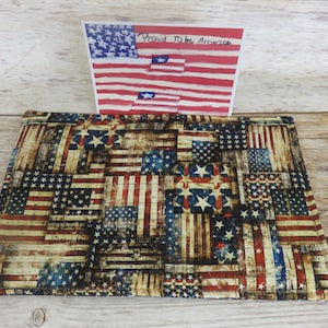 May include: Rectangular placemat with a patchwork design of the American flag in red, white, and blue. A small card with the American flag and the words "Proud to be American" is placed on top of the placemat.