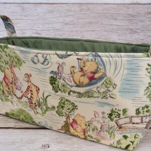 May include: A Winnie the Pooh themed fabric storage basket with green handles. The basket features a white background with illustrations of Winnie the Pooh, Piglet, Tigger, and Eeyore.