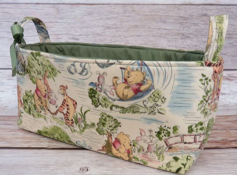 May include: A Winnie the Pooh themed fabric storage basket with green handles. The basket features a white background with illustrations of Winnie the Pooh, Piglet, Tigger, and Eeyore.